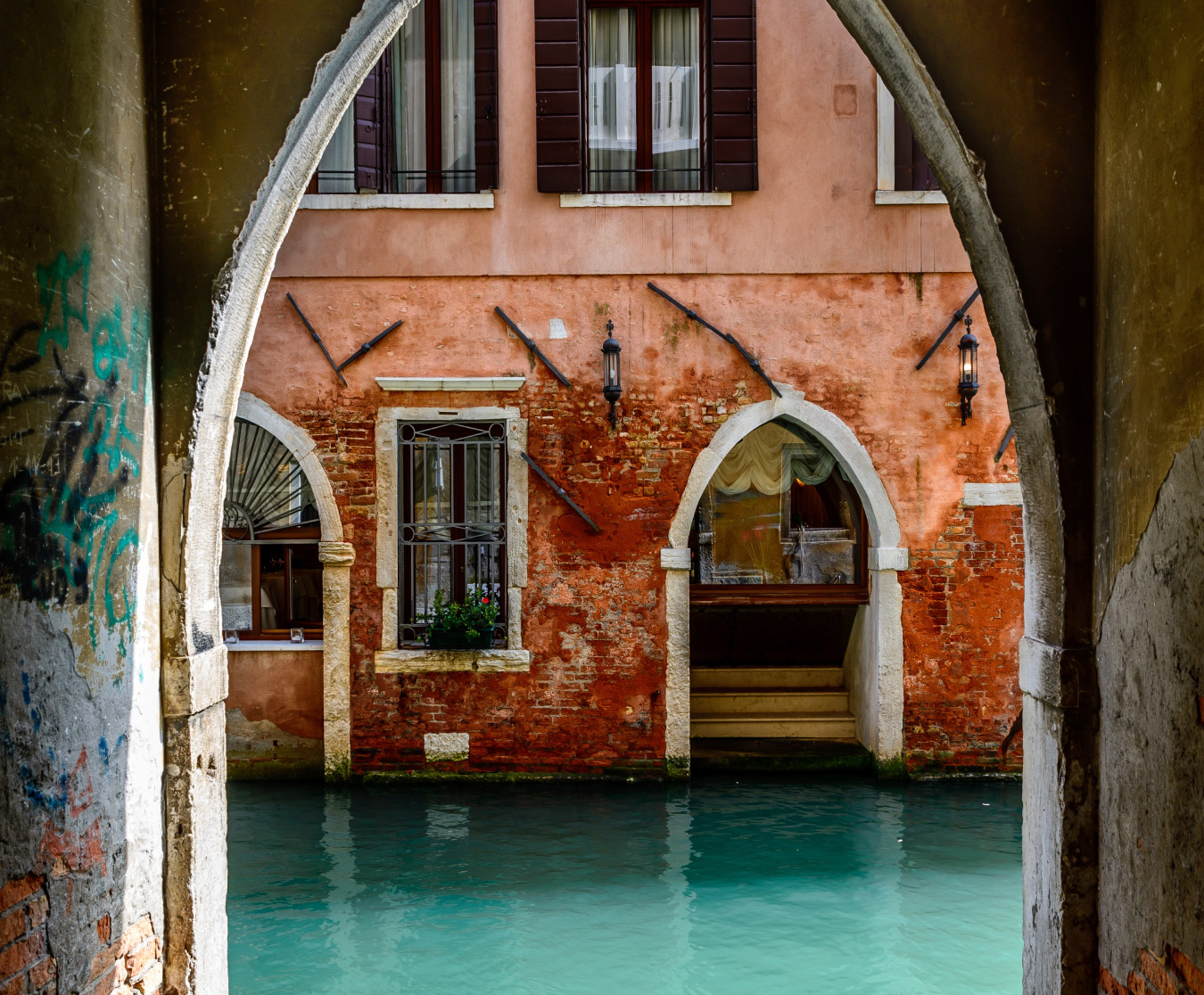 A venice scene showing a two story stucco building with arched doorways and exposed brick with a canal as seen through an arched doorway with graffiti on its walls