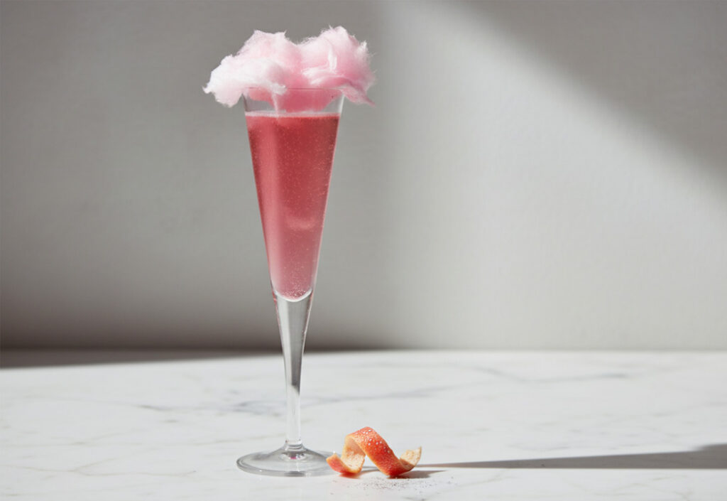 Close up of a pink cocktail in a champagne flute glass with fluffy pink cotton candy garnish on a marble surface